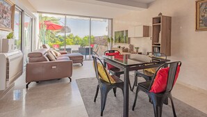 A modern living room with a glass dining table, leather sofa, and a view of a garden through large windows.