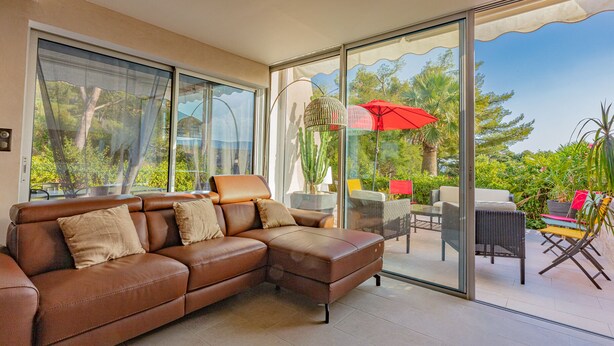 A modern living room with a brown leather sectional sofa, a glass door leading to an outdoor patio with a red umbrella, wicker furniture, and potted plants.