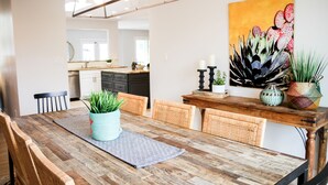 Un comedor con una mesa de madera, sillas de mimbre, un mantel tejido y un cuadro de flores en la pared.