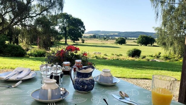 Une table dressée pour le petit-déjeuner, avec une vue panoramique sur un paysage verdoyant et des collines en arrière-plan.