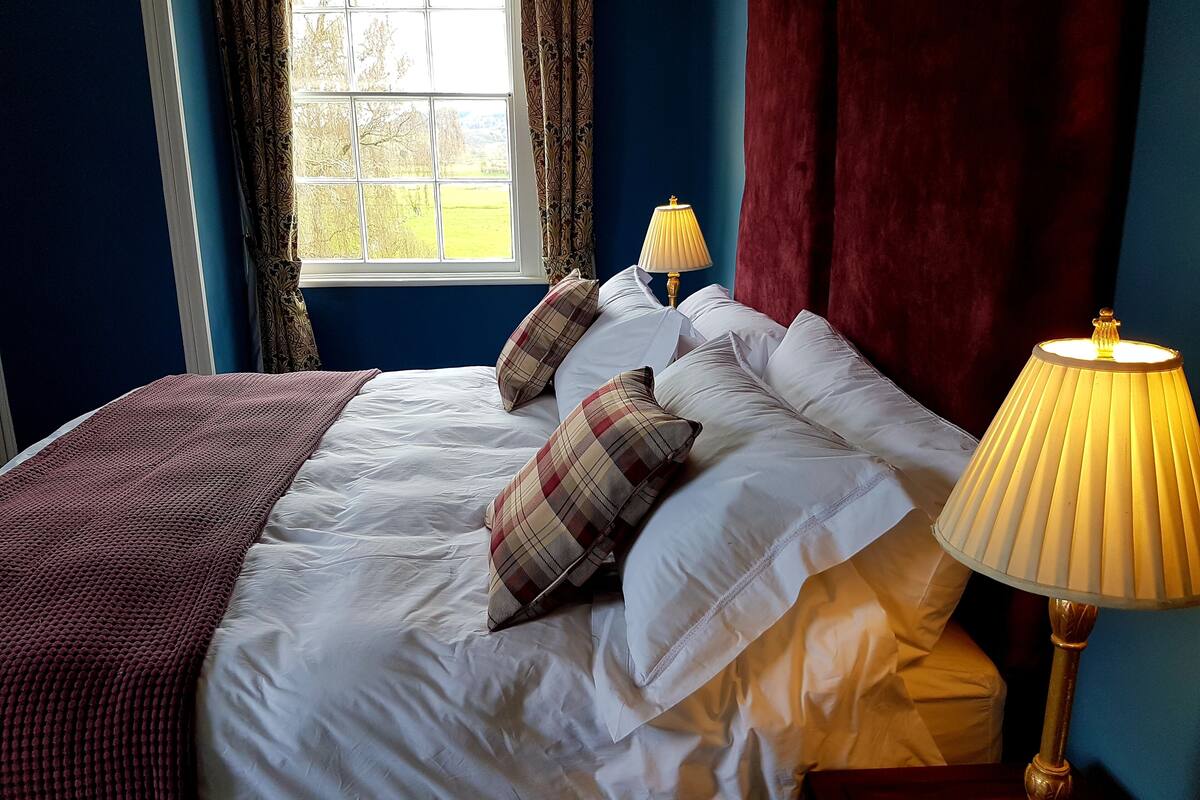 A neatly made bed with white linens and plaid pillows, a red throw blanket, and a bedside lamp.