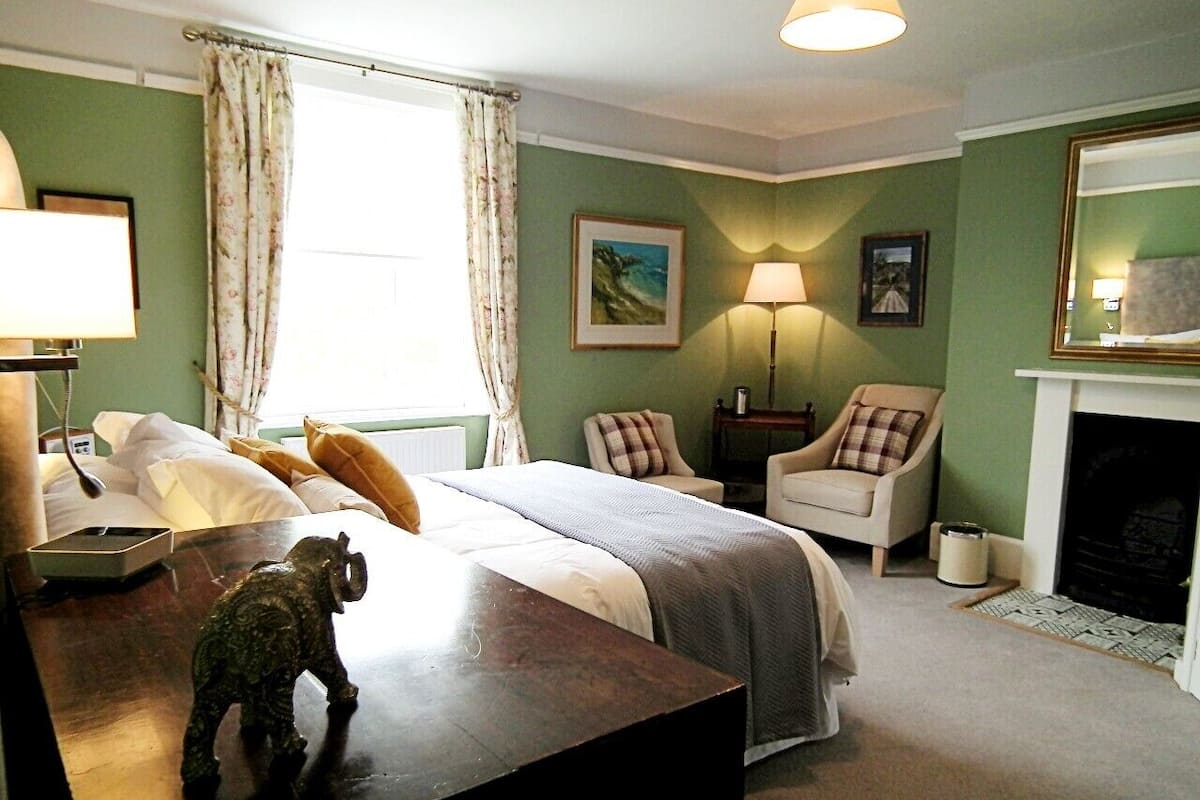 A neatly arranged bedroom with a bed, two chairs, a desk, a lamp, and a fireplace.