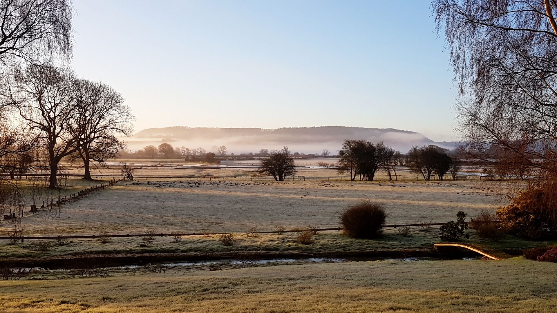 A rural landscape with a river, trees, and a distant mountain range.