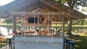 Ein überdachter Außenbereich mit Bar, einem Holzdeck, einem Pool im Hintergrund und einem Baum zur Rechten.