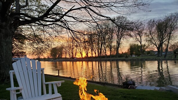 Ein Lagerfeuer mit Holzscheiten, ein weißer Adirondack-Sessel und ein See mit Bäumen im Hintergrund.