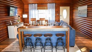 Interior de una cabaña con detalles de madera, una zona de bar con taburetes, un sofá, un televisor y un ventilador de techo.