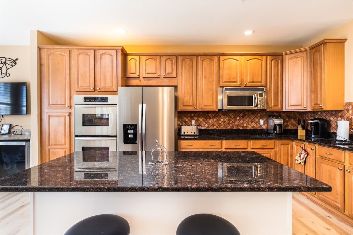 A kitchen with wooden cabinets, stainless steel appliances, a granite countertop, and a bar stool.