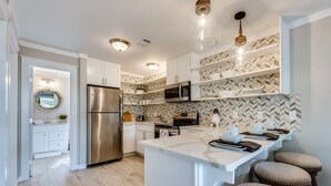 Una cocina moderna con armarios blancos, un refrigerador de acero inoxidable, un microondas y un salpicadero con estampado de espiga. Hay tres lámparas colgantes, una isla blanca con taburetes y un banco de madera.