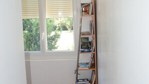 Un couloir avec un plancher en bois, une porte blanche, une fenêtre avec des stores, une horloge murale et une bibliothèque.