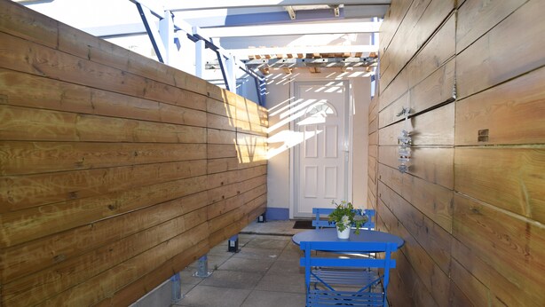 Un étroit couloir en bois, avec une table et des chaises bleues, une plante en pot et une porte au fond.