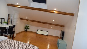 Une chambre spacieuse aménagée en mezzanine, avec un plafond en pente, des poutres en bois, une grande fenêtre, un lit avec une literie à motifs, une table de chevet, une chaise et une plante en pot.