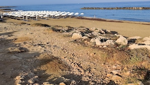 Ein Strand mit weißen Sonnenschirmen und Liegestühlen, felsigem Untergrund und blauem, klarem Himmel.