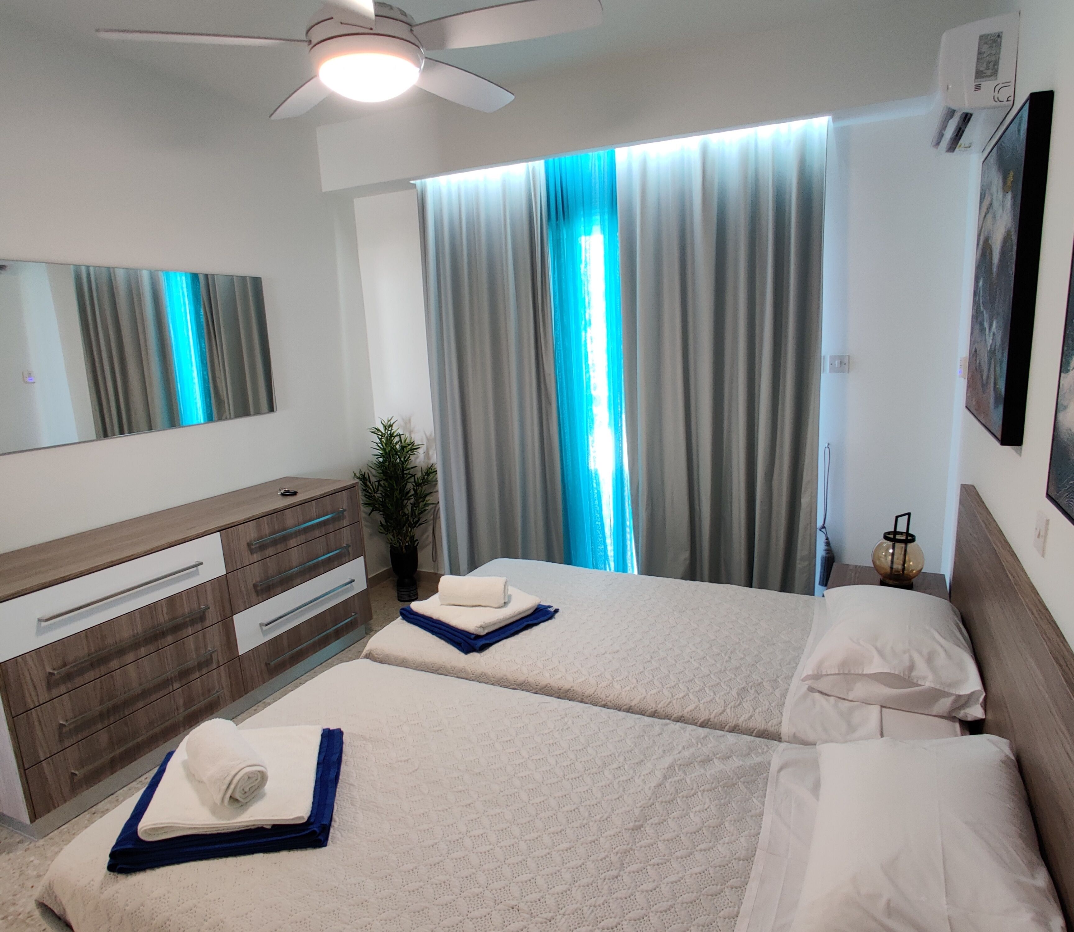 A hotel room with two beds, a large mirror, a ceiling fan, and a window with curtains.