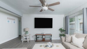 A living room with a ceiling fan, a flat-screen TV, a coffee table with decorative items, a sofa, and a side table with a lamp. - New updated 1.5 blocks to beach, 5 min walk, 4 bedroom 3 bath, king size bed,  (Galveston)