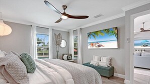A bedroom with a bed, a ceiling fan, a painting of a beach scene, and a view of the outdoors through a window.