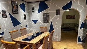 A dining area with a wooden table and wicker chairs, a geometric-patterned wall, and a sofa in the background.