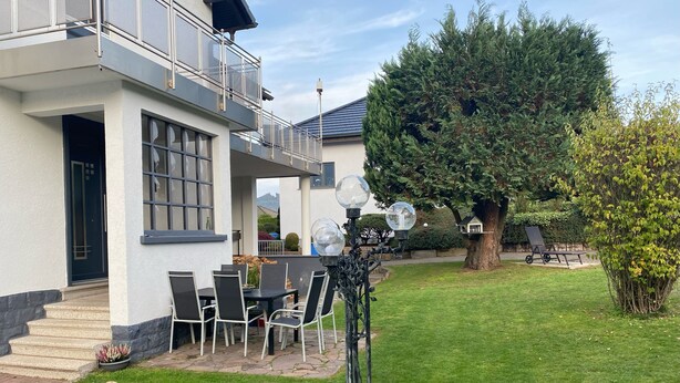 A modern house with a balcony, a dining area with chairs, and a well-maintained lawn.