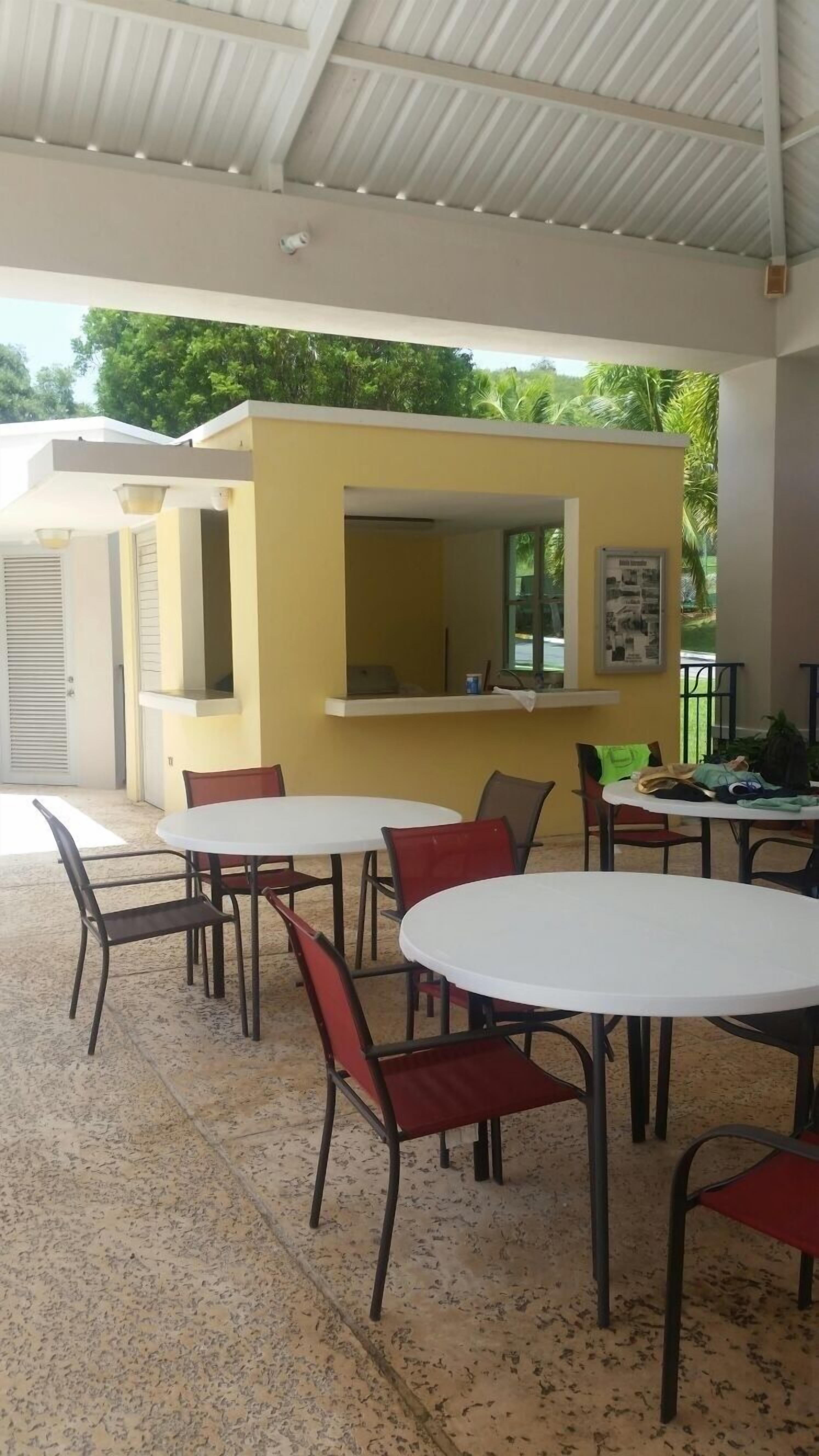 An outdoor cafe area with round tables and chairs, a yellow counter, and a view of greenery.