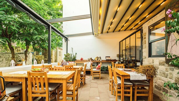 Un patio al aire libre con mesas y sillas de madera, muros de piedra y plantas en macetas.