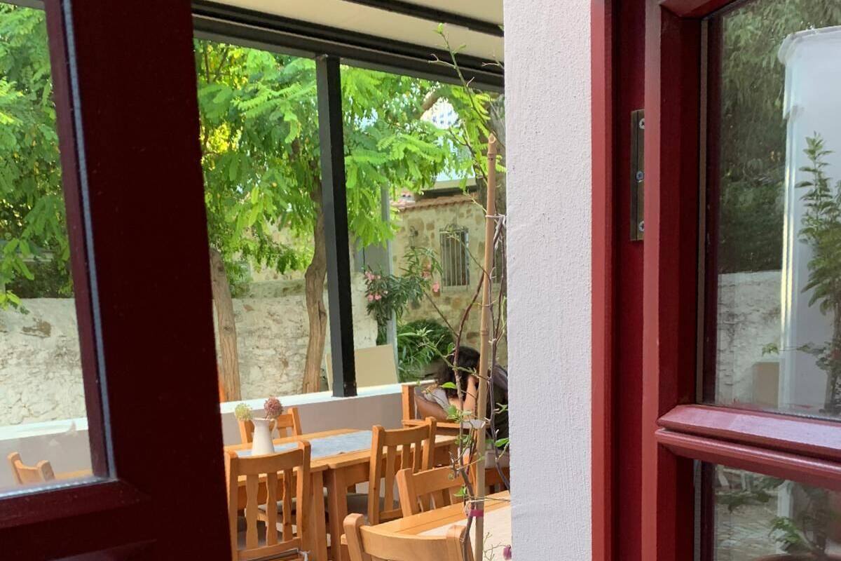A view through a window of a patio area with wooden tables and chairs, a potted plant, and a stone wall in the background.