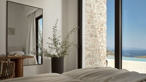 Une chambre à coucher avec un lit, une table de chevet sur laquelle repose un livre, une plante en pot et une grande fenêtre donnant sur un mur en pierre et offrant une vue sur la mer.