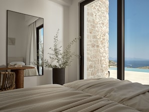 Une chambre à coucher avec un lit, une table de chevet sur laquelle repose un livre, une plante en pot et une grande fenêtre donnant sur un mur en pierre et offrant une vue sur la mer.