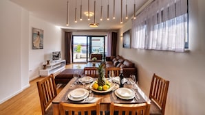A dining area with a wooden table set for four, surrounded by wooden chairs. A fruit platter with a pineapple, lemons, and limes is placed in the center. The room has a modern design with a large window covered by sheer curtains, a brown leather sofa, and a white wall with a painting. - Deluxe Apartment Isabela Prague (Zadar)