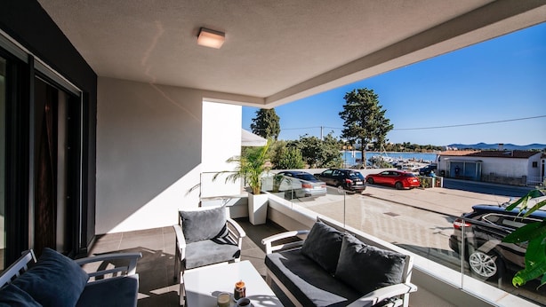 A balcony with a sofa, table, and chairs, overlooking a parking area and a marina. - Deluxe Apartment Isabela Prague (Zadar)