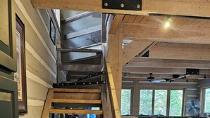 Eine Holztreppe mit einem Maschendrahtgeländer führt zu einem Küchenbereich mit einem großen Fenster und einem Ventilator.