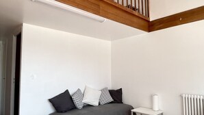 A room with a wooden staircase, a grey sofa with patterned cushions, a small round table, and a radiator. - Studio in a magnificent villa with private beach access. (Esquibien)