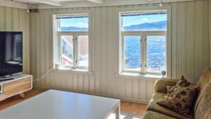 Een woonkamer met een televisie, een witte salontafel, een groene bank en twee ramen met uitzicht op zee en bergen.