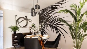 Uma sala de jantar com uma mesa redonda posta para quatro pessoas, rodeada por cadeiras pretas com pés de madeira. Há duas luminárias pendentes sobre a mesa.