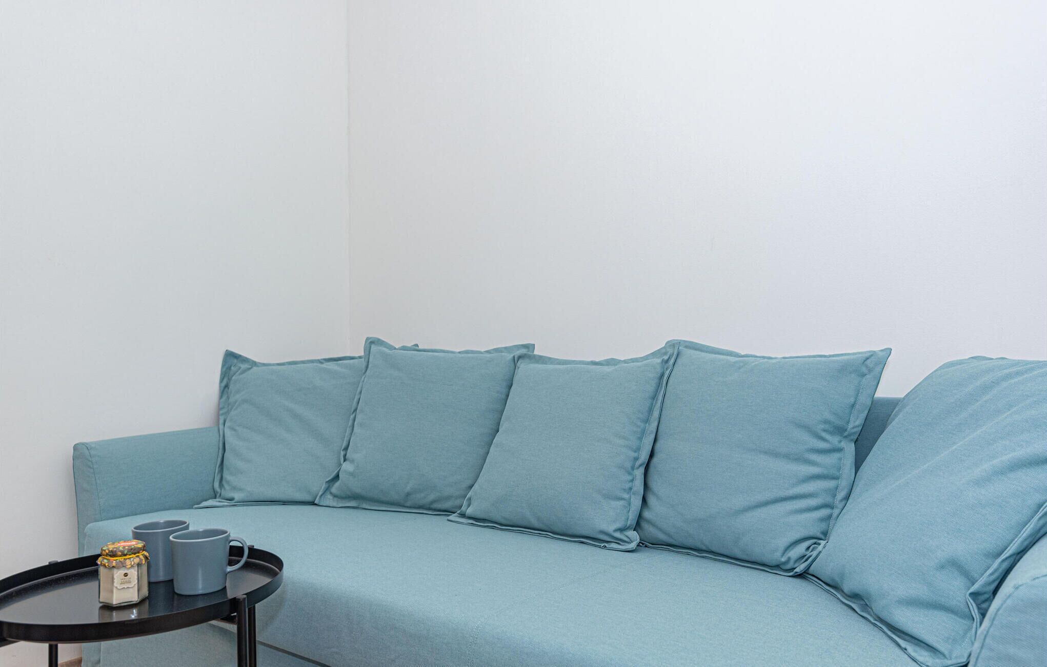 Um sofá azul claro com quatro almofadas, uma mesa redonda pequena com uma bandeja contendo duas canecas cinzas e um objeto decorativo, em frente a uma parede branca lisa.