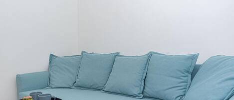 Um sofá azul claro com quatro almofadas, uma mesa redonda pequena com uma bandeja contendo duas canecas cinzas e um objeto decorativo, em frente a uma parede branca lisa.