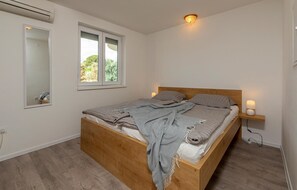 A bedroom with a wooden bed, a window with a view of greenery, a wall-mounted air conditioner, and a bedside table with a lamp. - Beautiful home in Rosini (Rosini)