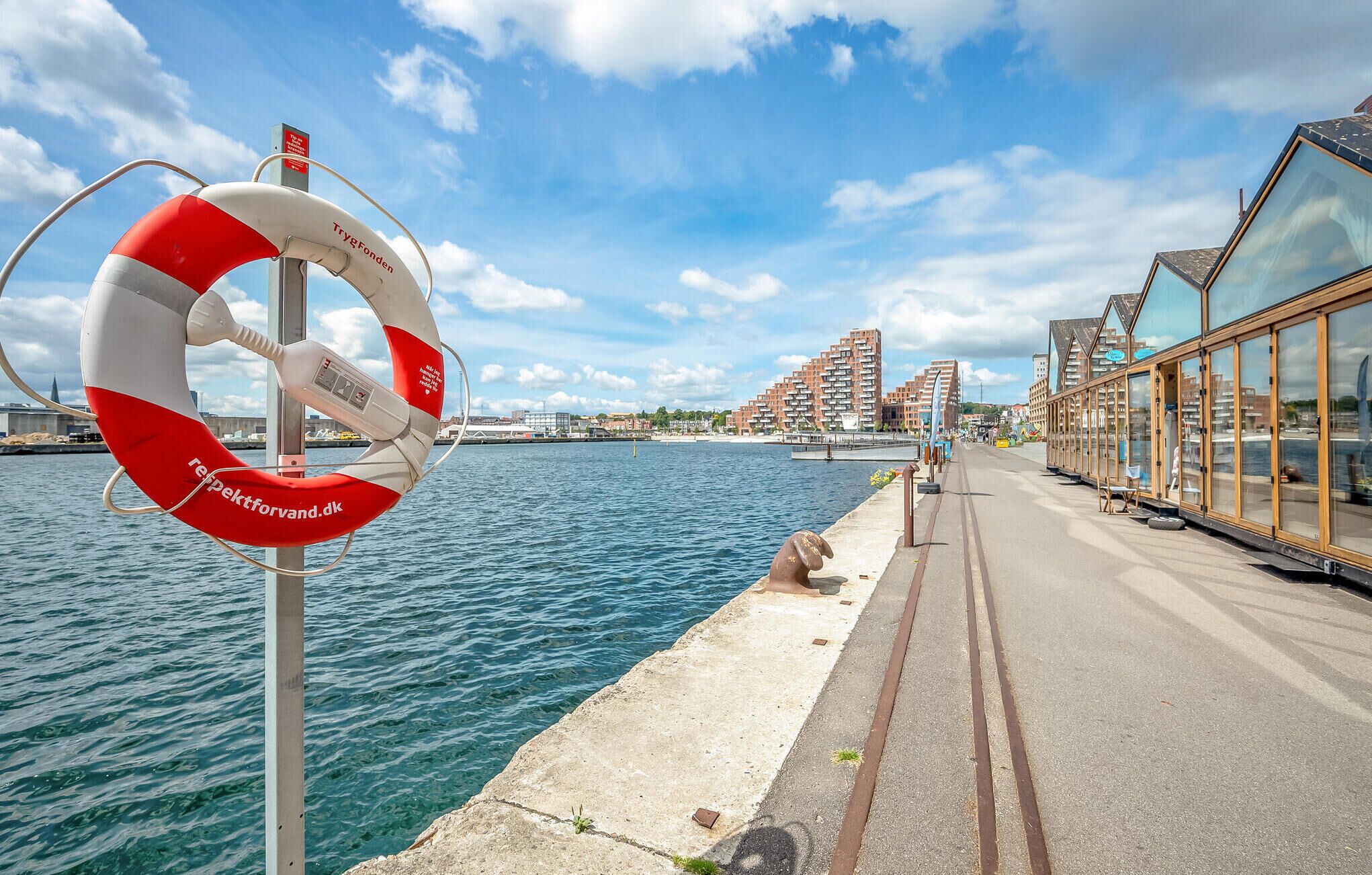 Eine Uferpromenade mit einem Rettungsring, einem Betonweg und modernen Gebäuden im Hintergrund.