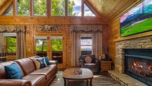 A cozy cabin interior with a leather sofa, a stone fireplace, and a large TV screen displaying a scenic landscape. - Sunset Mountain Views • 3 King Suites • Hot Tub • Game Room • 8 mi to Dollywood (Sevierville)