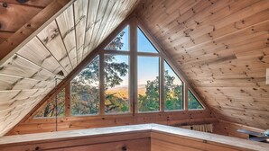 A wooden attic bedroom with large windows, a sloped ceiling, and a view of trees and hills. - Sunset Mountain Views • 3 King Suites • Hot Tub • Game Room • 8 mi to Dollywood (Sevierville)