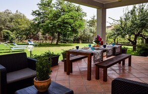 Ein möblierter Außenbereich mit überdachter Sitzgruppe, Tischen und Stühlen, umgeben von einem üppigen Garten und einem Pool.