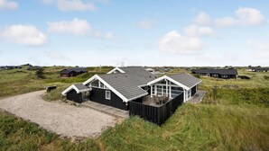 A coastal house with a gravel driveway, surrounded by grassy dunes and other similar houses in the background. - 4 bedroom stunning home in Hirtshals (Hirtshals)