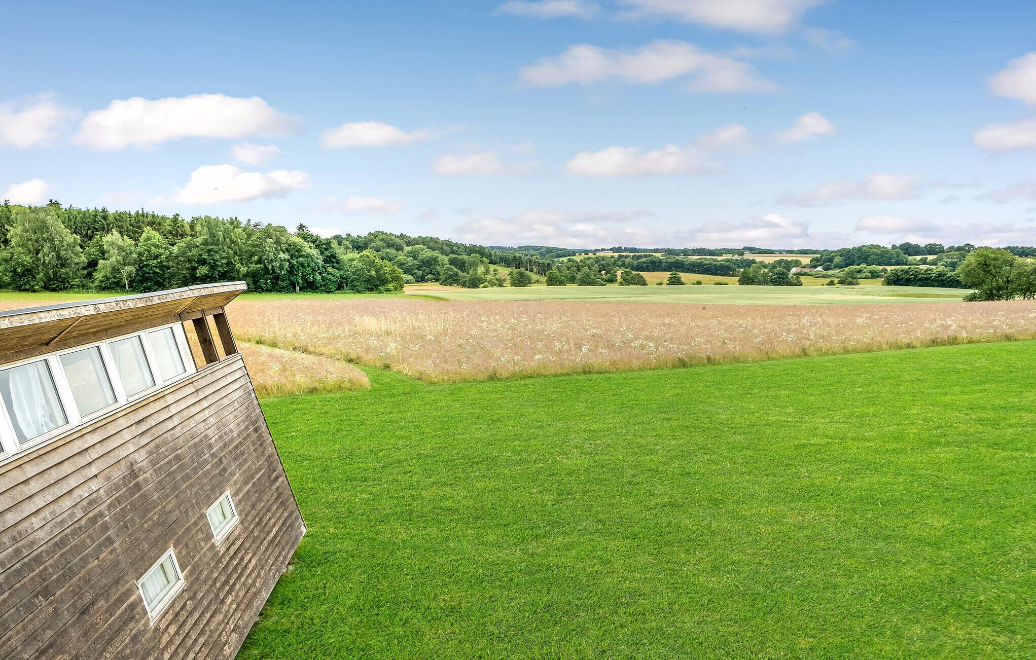 Ein Holzhaus mit großen Fenstern, die auf ein grünes Feld und eine entfernte Baumlinie unter blauem Himmel blicken.