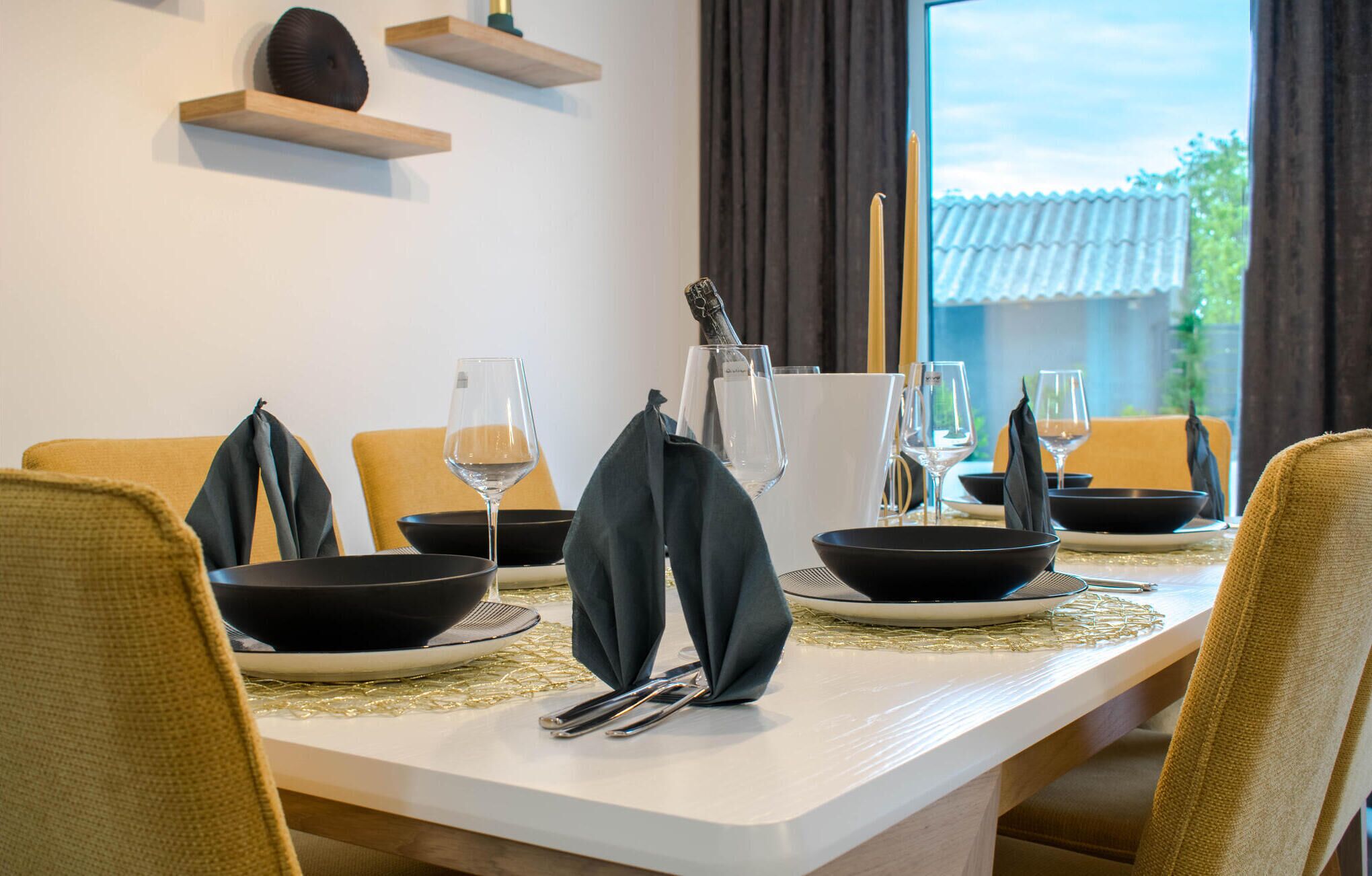 Een eettafel gedekt met zwarte kommen, wijnglazen en een fles water. Er staan stoelen rondom de tafel en op de achtergrond is een raam met gordijnen te zien.