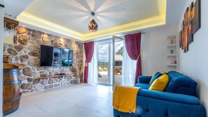 A living room with a stone wall, a flat-screen TV, a blue sofa with yellow cushions, and a wooden barrel. - Amazing home in Vrlika with house a mountain view (Vrlika)