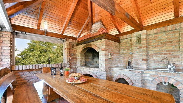 Cocina rústica al aire libre con una mesa de madera, horno de ladrillo y vistas a la vegetación.