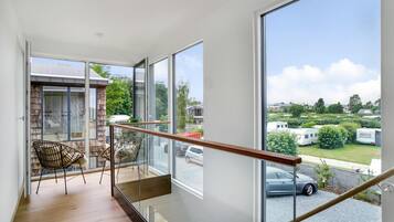 A spacious balcony with a wooden floor, large glass doors leading to a garden, and a view of a residential area with parked cars and greenery.