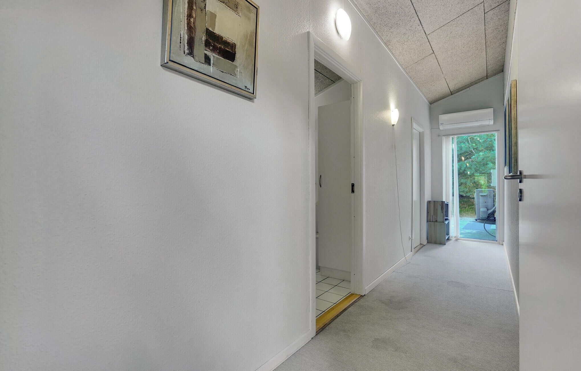 A hallway with white walls, a door leading to a room, and a view of a garden through a glass door.
