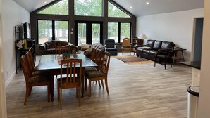 Amplia sala de estar con techo alto, ventanas grandes, una mesa de comedor con sillas, un sofá, un sillón de cuero y una lámpara.