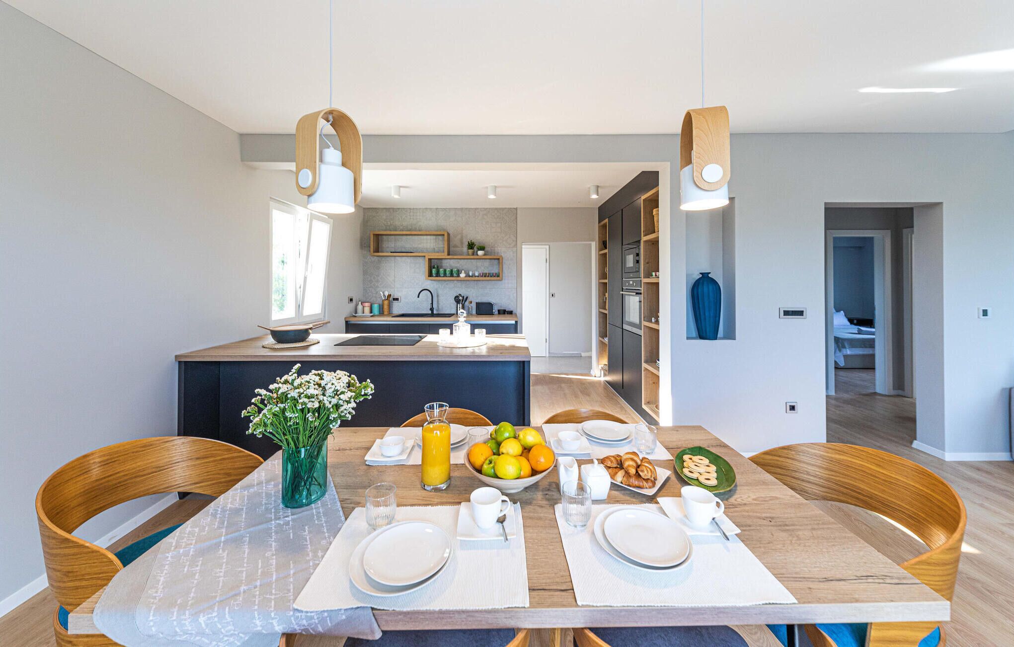 Una cucina moderna con isola centrale, sedie di legno, tavolo da pranzo apparecchiato per la colazione e vista sull'area soggiorno adiacente.