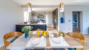 A modern kitchen with a central island, wooden chairs, a dining table set for breakfast, and a view into the adjacent living area. - Lovely home in Kastel Stafilic (Kastel Stafilic)