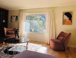 A living room with a large window, two armchairs, a round table, and a bookshelf.